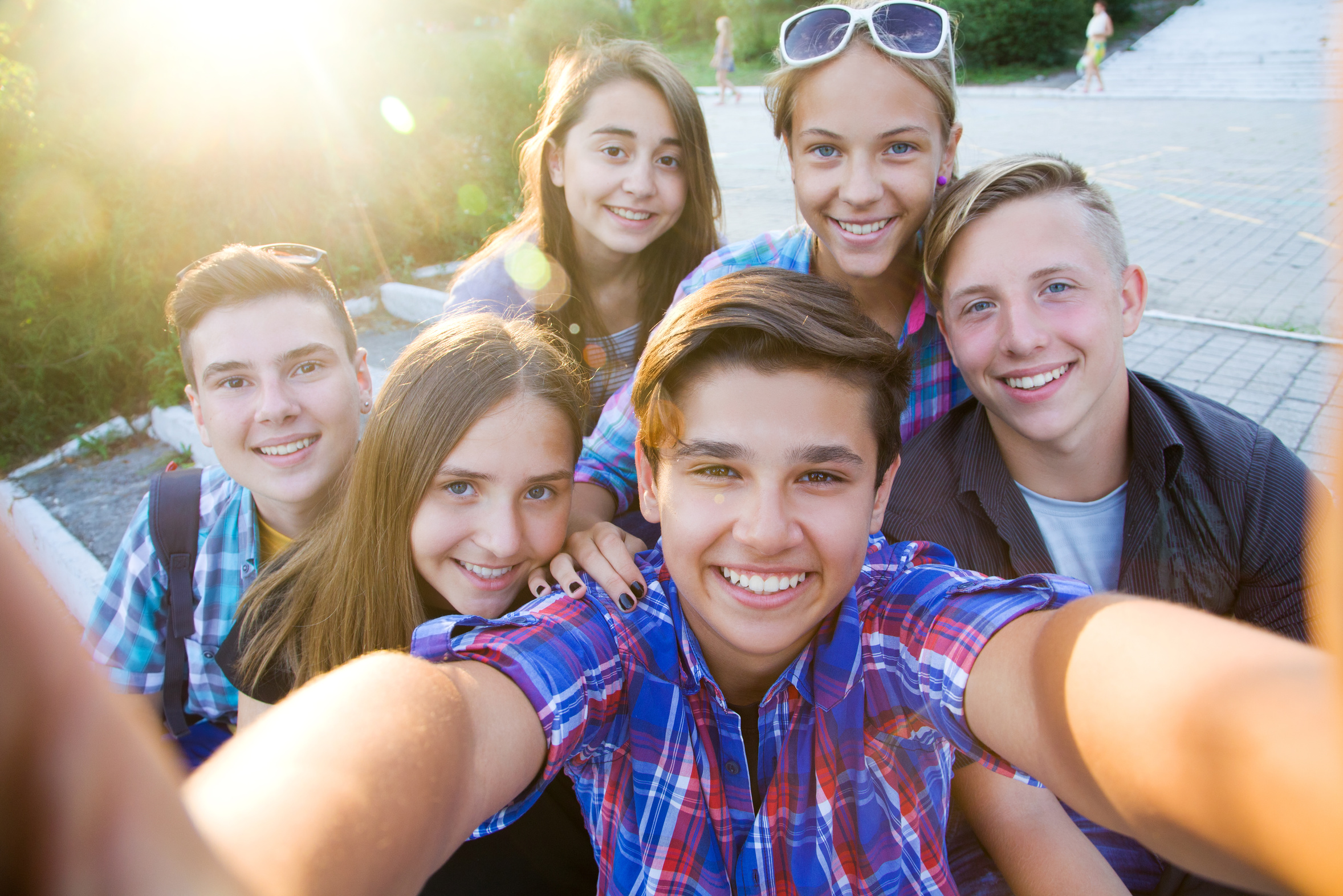 Ein dunkelhaariger Junge fotografiert sich mit beiden weit ausgestreckten Armen selbst. Um ihn herum sind drei Mädchen und zwei Jungen zu sehen, die alle in die Kamera lächeln. Im Hintergrund ist eine Grünanlage mit Personen in Freizeitkleidung zu sehen. Die linke obere Ecke des Bildes wird durch das Gegenlicht der Sonne komplett überstrahlt.