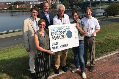 Fünf Männer und eine Frau stehen als Gruppe auf einer Rasenfläche am Südstrand und präsentieren eine große Tafel mit dem Aufdruck "European Energy Award". Im Hintergrund sind Hafen, Bontekai und die KW-Brücke zu erkennen.