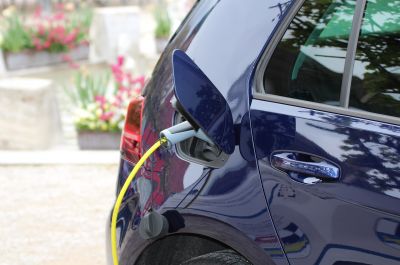 Das Heck eines dunkelblauen Autos mit einer geöffneten kleinen Klappe, in die ein Ladekabel gesteckt ist. Im unscharfen Hintergrund sind Blumenbeete zu erkennen. 