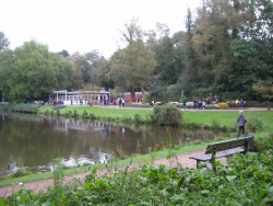 Blick über die Ecke des Teiches hinweg zum Bootshaus im Hintergrund.  Auf dem Weg vor dem Bootshaus viele Kinder und Jugendliche, teilweise in Sportkleidung.