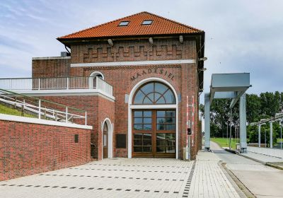 Klinkergebäude mit der Aufschrift "Maadesiel", am linken Bildrand der Deich, rechts eine Fahrbahn, die zum hinteren Bildrand über eine Hubbrücke führt.