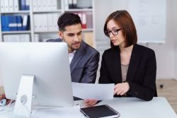 Eine junge Frau im Business-Kostüm und ein junger Mann im Anzug sitzen in einem modernen Büro am Schreibzisch. Sie zeigt ihm einen Zettel mit Informationen, über die sie sich offenbar unterhalten.