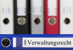 Im Vordergrund liegt ein blauer Aktenordner mit der Aufschrift "Verwaltungsrecht". Im Hintergrund ist unscharf eine Reihe weiterer Aktenordner zu erkennen.