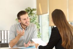 Eine junge Frau im Businesskostüm ist schräg von hinten am Schreibtisch zu sehen. In der Hand hält sie einen Zettel. Auf der anderen Seite des Tisches sitzt ihr zugewandt ein lächelnder junger Mann. Beide unterhalten sich offenbar angeregt.