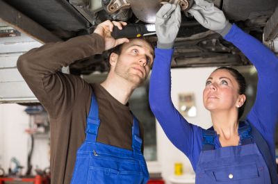 Eine junge Frau und ein junger Mann in blauen Latzhosen arbeiten  konzentriert an der Unterseite eines Autos, das auf einer Hubbühne angehoben wurde.