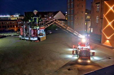 Drehleiterwagen auf dem Hof der Feuerwehr, aufgenommen bei Dunkelheit aus der Luft. Im Vordergrund der Korb der ausgefahrenen Drehleiter mit einem Feuerwehrmann.