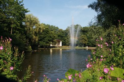 Blick über einen Teich zwischen blühenden Pflanzen hindurch. Mitten im Teich eine Fontäne, an der sich ein Regenbogen bildet. Dahinter eine halbkreisförmige Bühne mit einem überstehenden Dach.