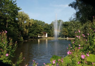 Blick über einen Teich zwischen blühenden Pflanzen hindurch. Mitten im Teich eine Fontäne, an der sich ein Regenbogen bildet. Dahinter eine halbkreisförmige Bühne mit einem überstehenden Dach.