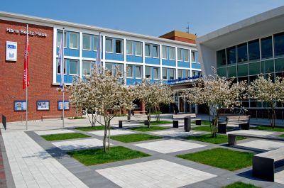 VHS-Gebäude und Stadtbibliothek, davor die schachbrettartige Anordnung von gepflasterten und grünen Flächen. In den grünen Flächen stehen kleine Bäume, die weiß blühen.