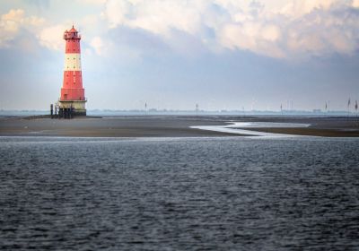 Der rote Leuchtturm Arngast mit seinem breiten weißen Querstreifen ist links im oberen Bilddrittel abgebildet. Im Vordergrund sind die Wellen des Meeres zu erkennen, rund um den Leuchtturm im Hintergrund eine bei Ebbe trockengefallene Sandbank.