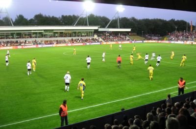 Ein Fußballspiel unter Flutlicht im Jadestadion. Im Vordergrund sind die Köpfe der Zuschauer auf der vollbesetzten Tribüne zu sehen.