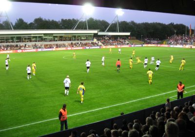 Ein Fußballspiel unter Flutlicht im Jadestadion. Im Vordergrund sind die Köpfe der Zuschauer auf der vollbesetzten Tribüne zu sehen.